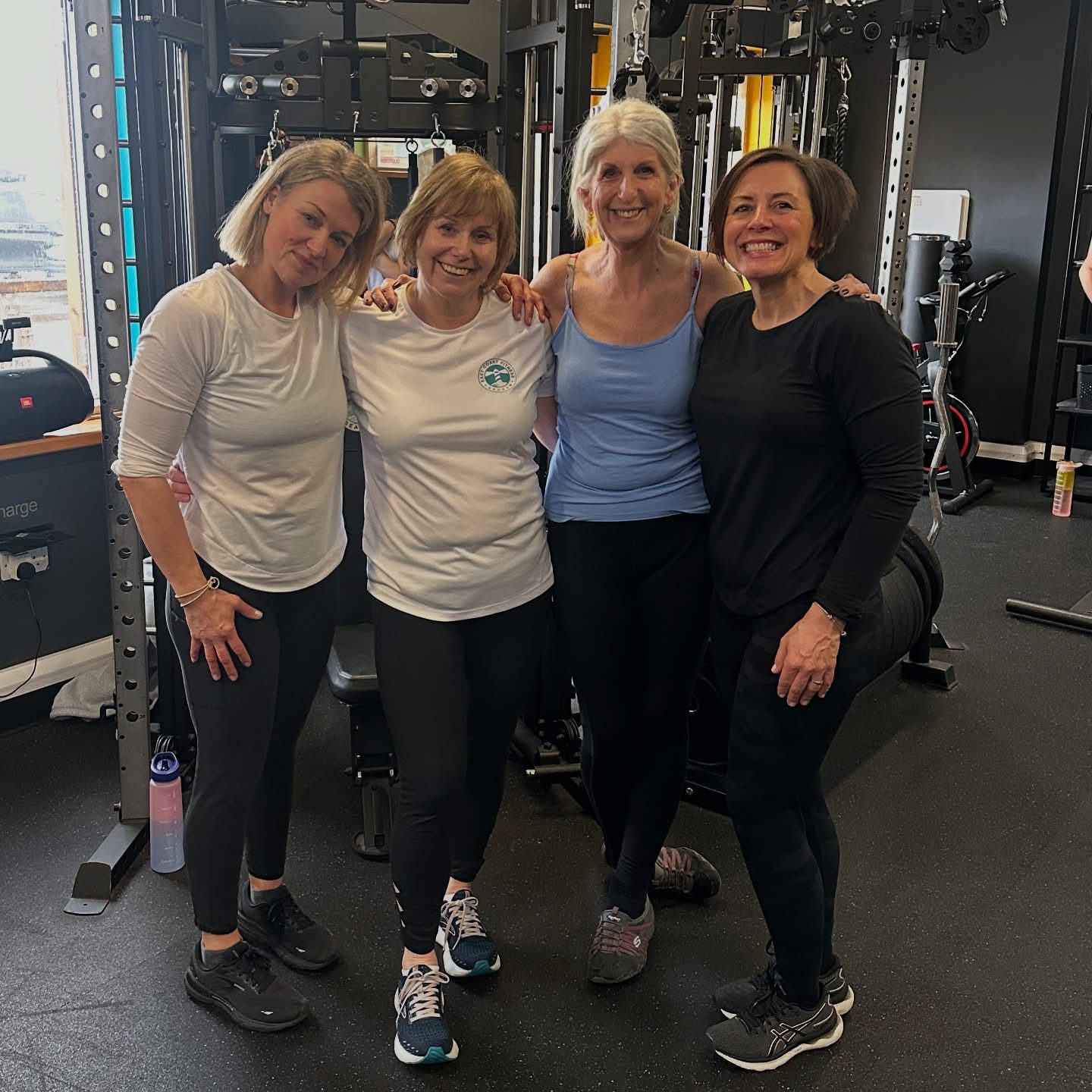 Four women smiling and posing together in a gym setting, wearing athletic clothing, showcasing a supportive fitness community.