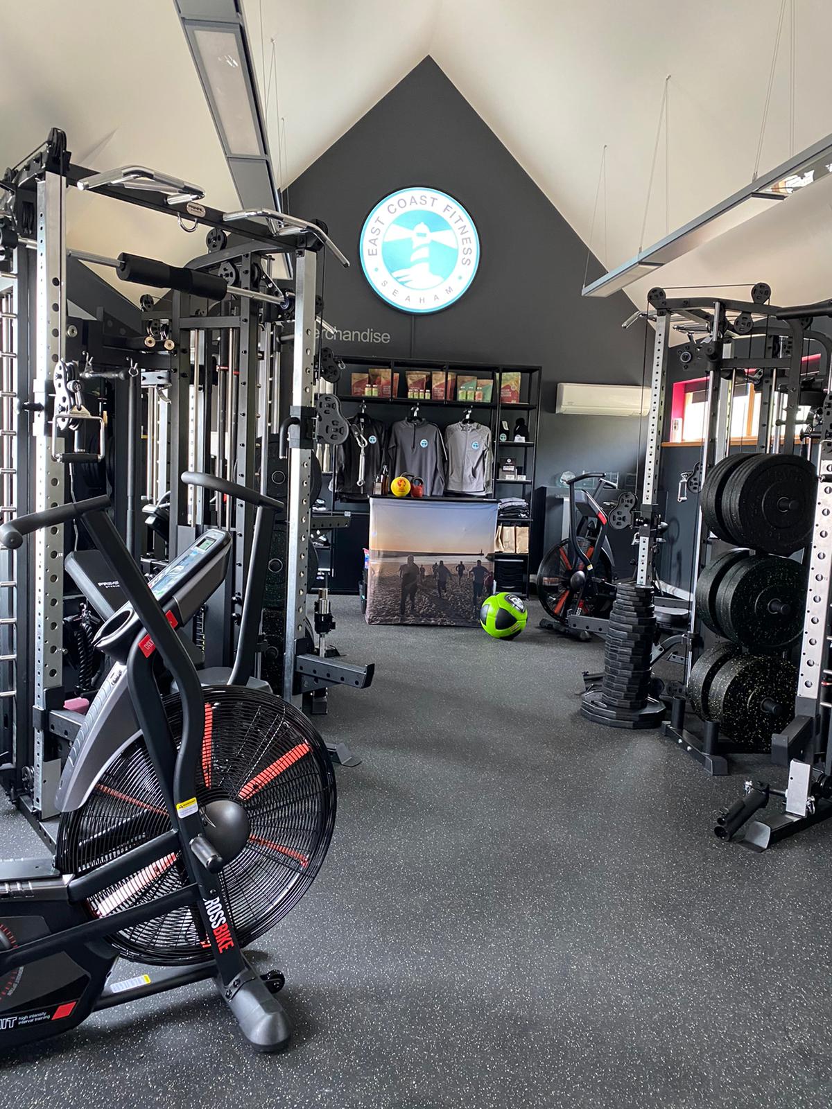 Interior view of East Coast Fitness gym featuring various workout equipment, including an air bike, weightlifting racks, and exercise balls, with a merchandise display in the background.