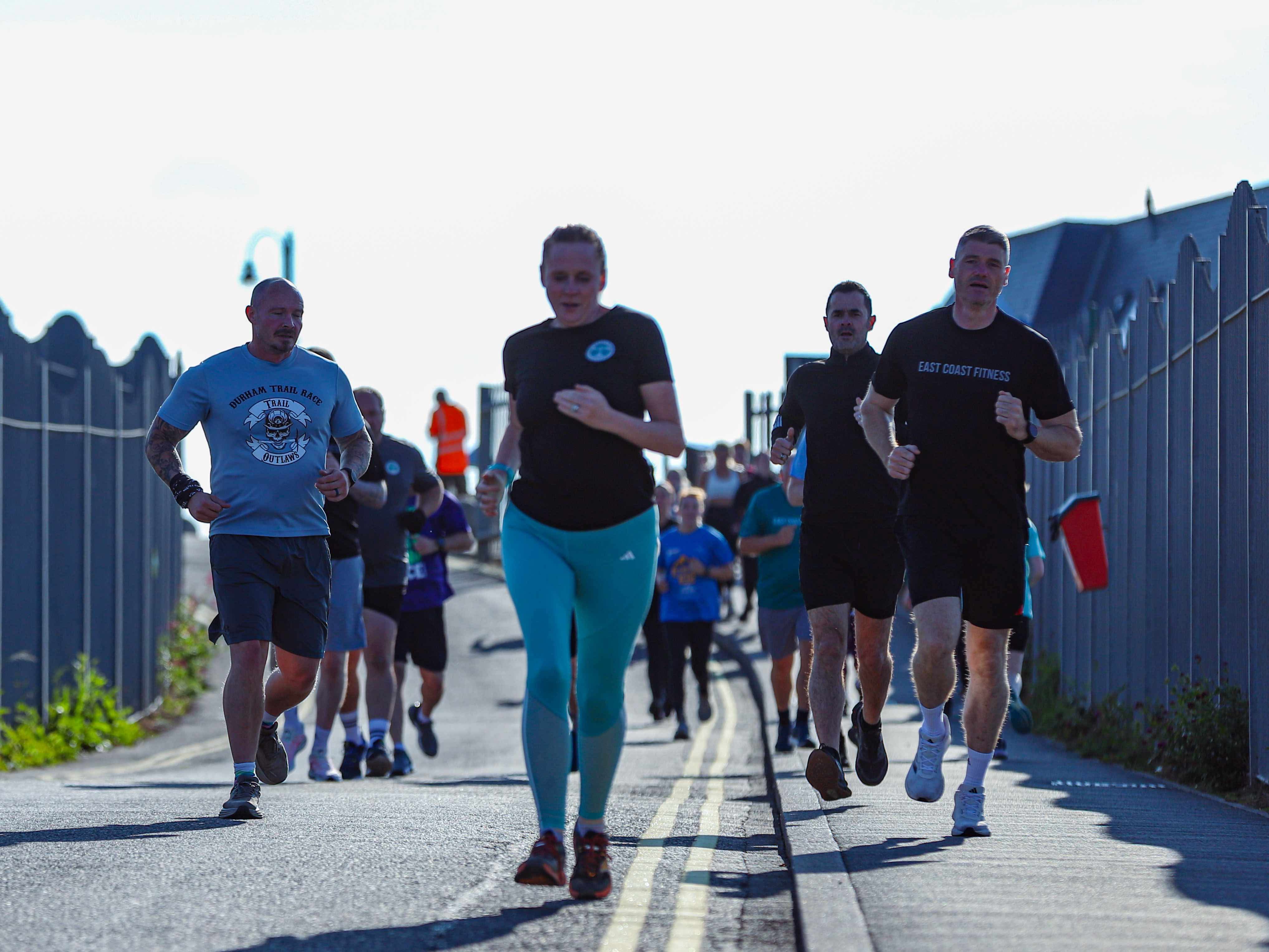 A group of diverse people jogging along a path, some wearing fitness attire, with a fence in the background under clear skies.