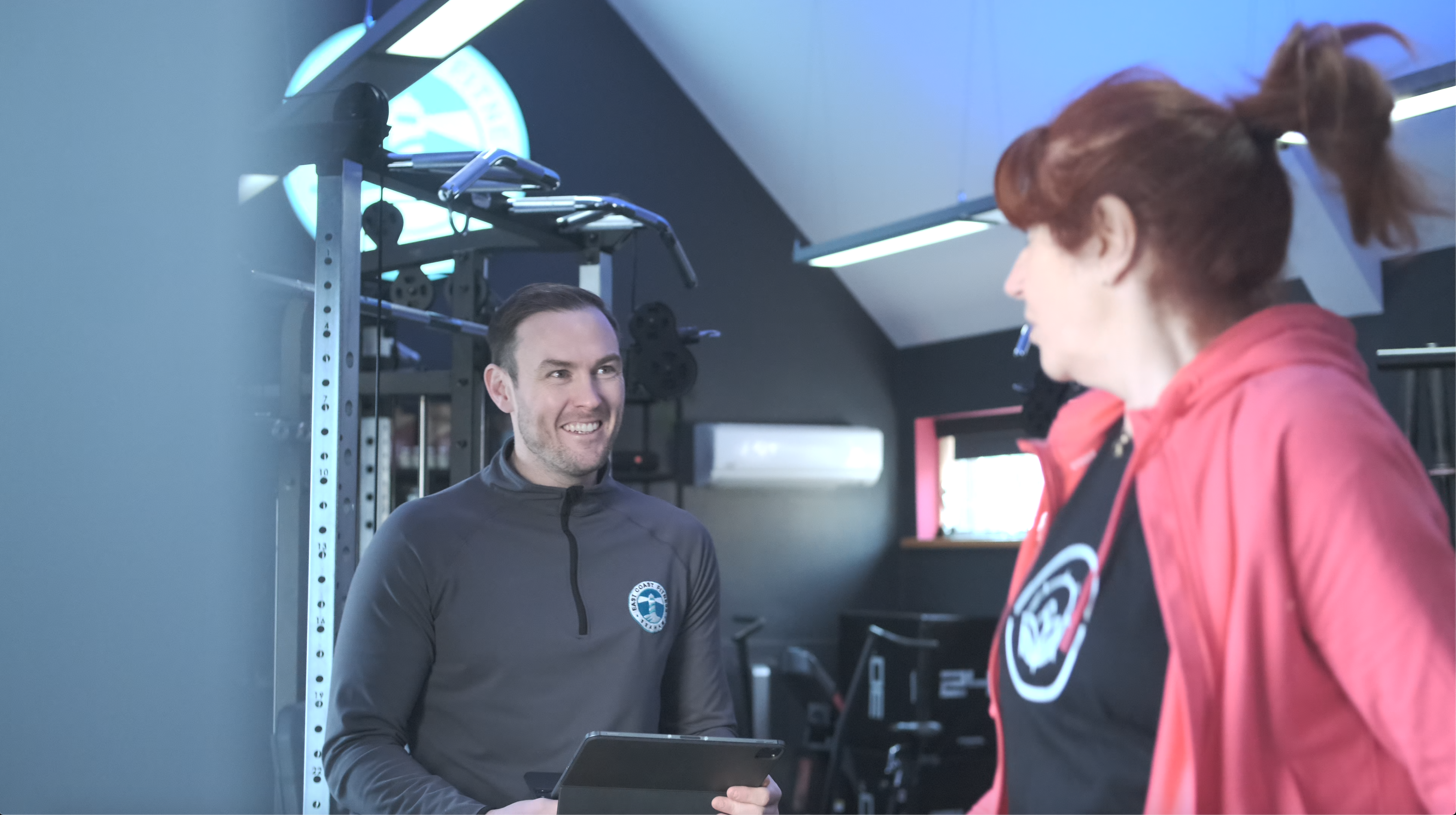 A fitness instructor smiling and holding a tablet while discussing with a woman in a gym setting.
