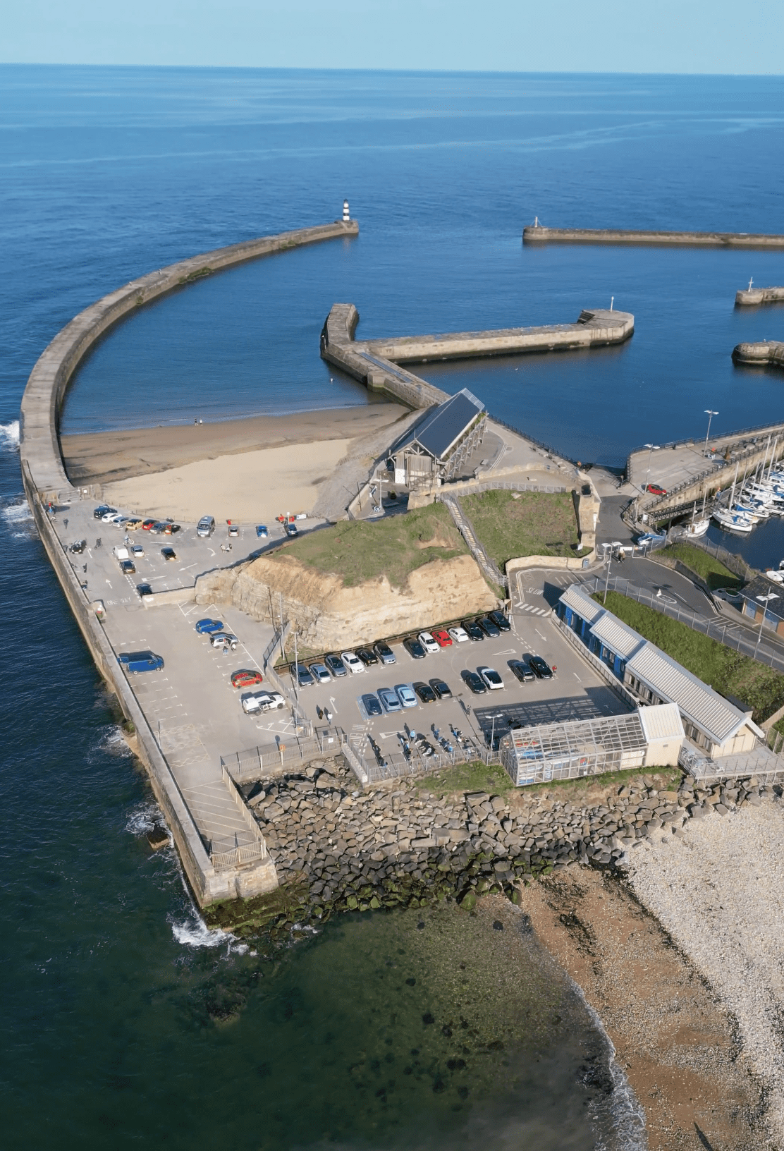 Aerial view of a coastal area featuring a curved pier, boats, and a parking lot near the beach.