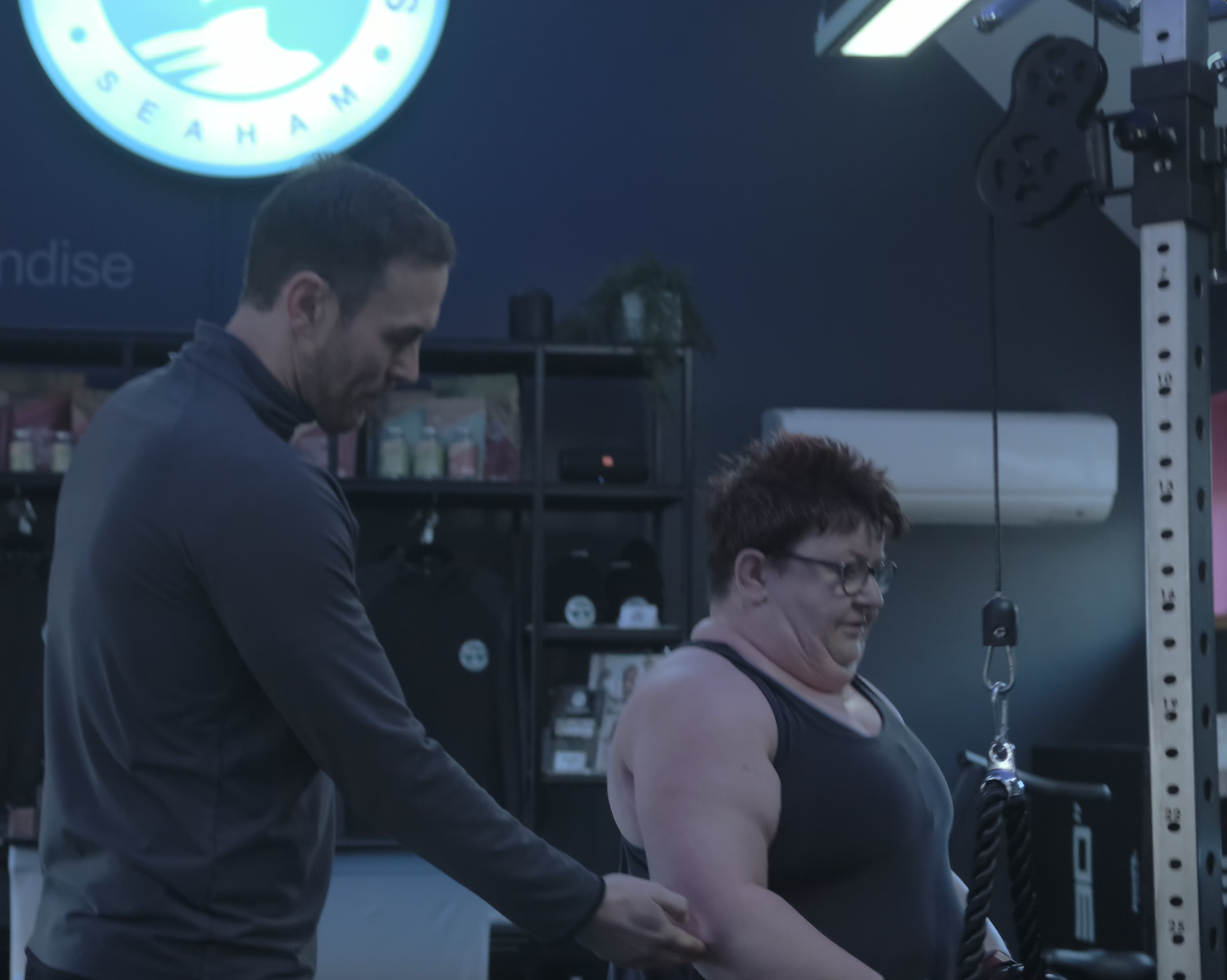 A trainer assists a participant during a workout session at a gym, with fitness equipment and merchandise in the background.