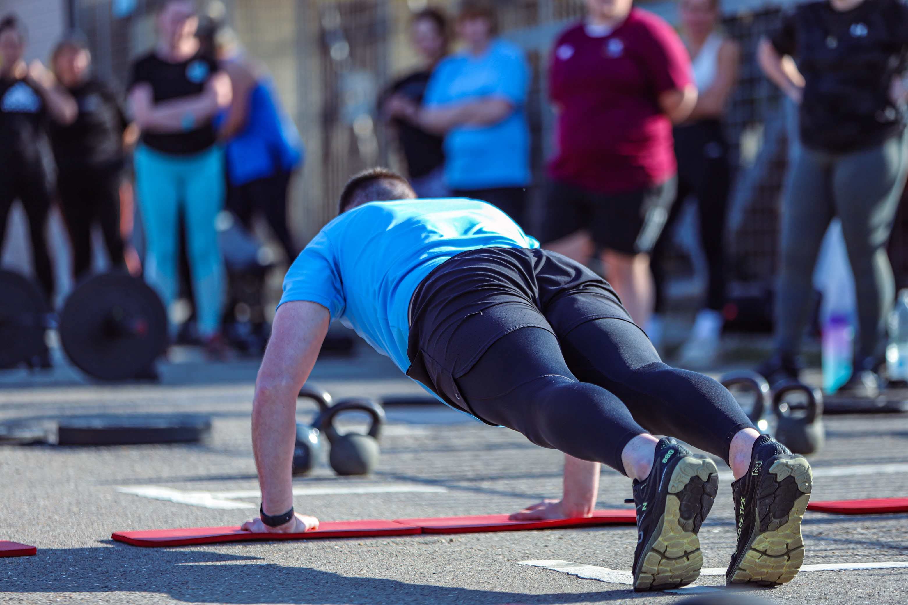 A person performing push-ups in an outdoor fitness class, surrounded by others watching, on a clear day.