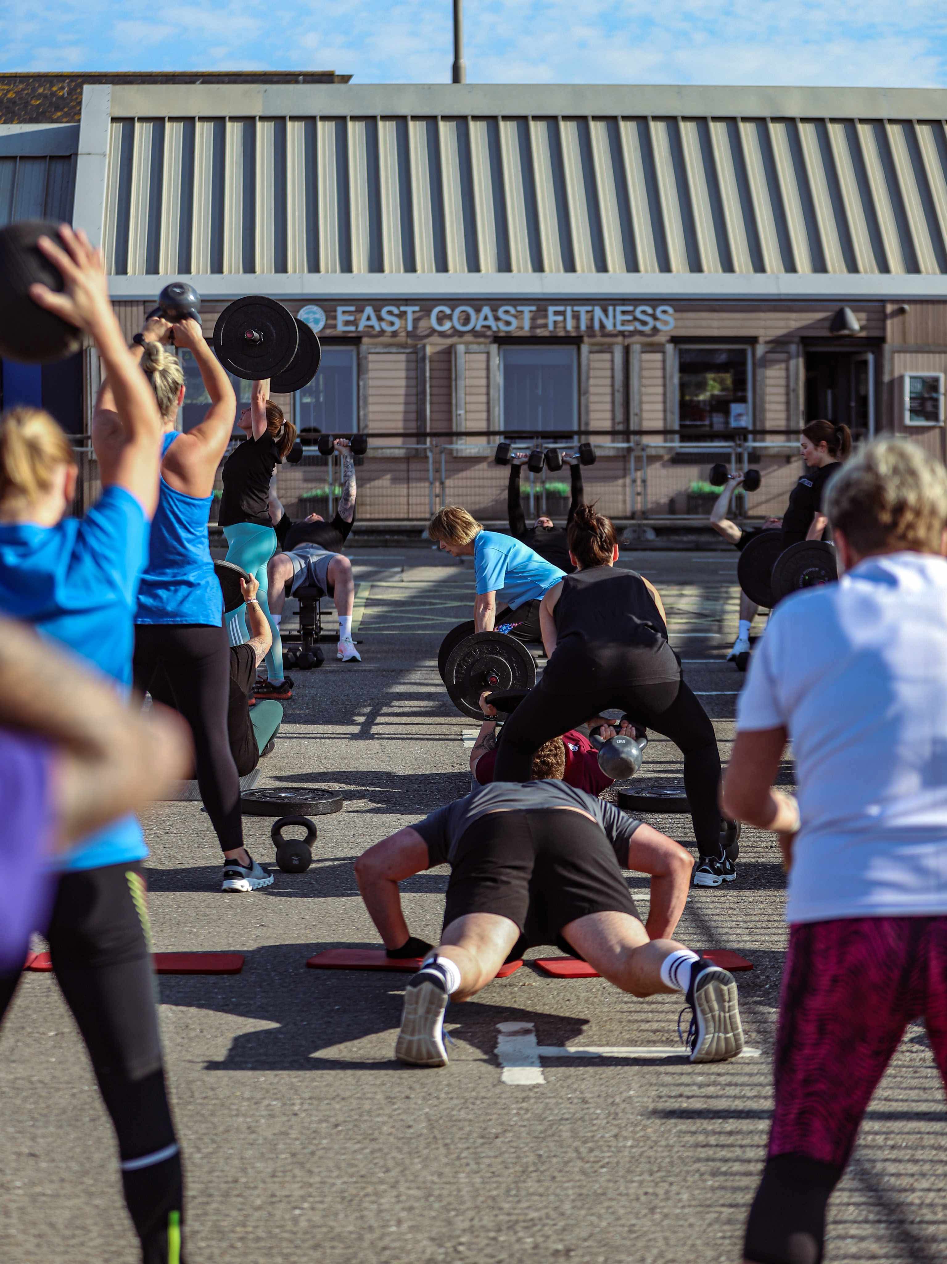 A group of people participating in an outdoor fitness session at East Coast Fitness, engaging in various exercises with weights.